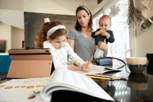 Mother multitasking with baby and daughter doing homework at the table.