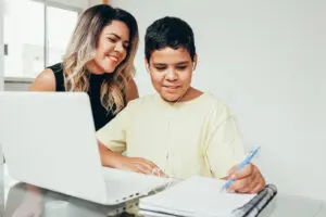 A woman smiles at a boy wearing headphones while he writes in a notebook, with a laptop in front of them.